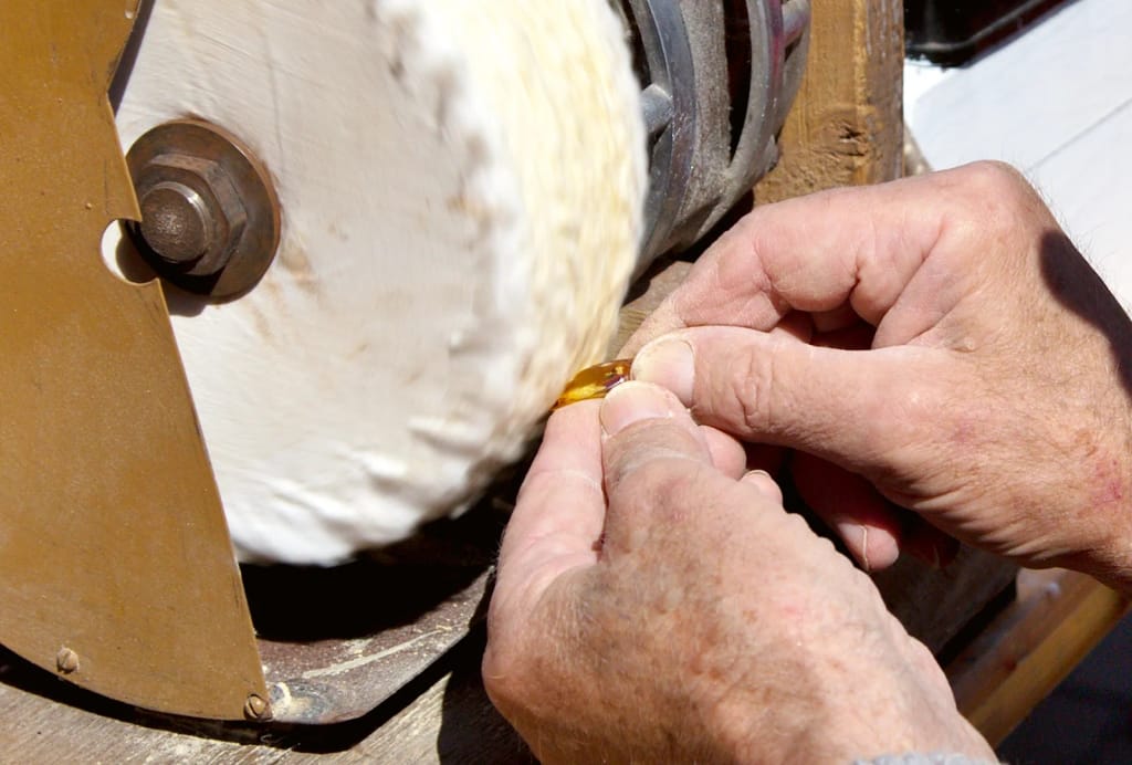 Hands polish a small amber stone on a spinning buffing wheel. The workshop scene highlights precision, care, and fine craftsmanship.